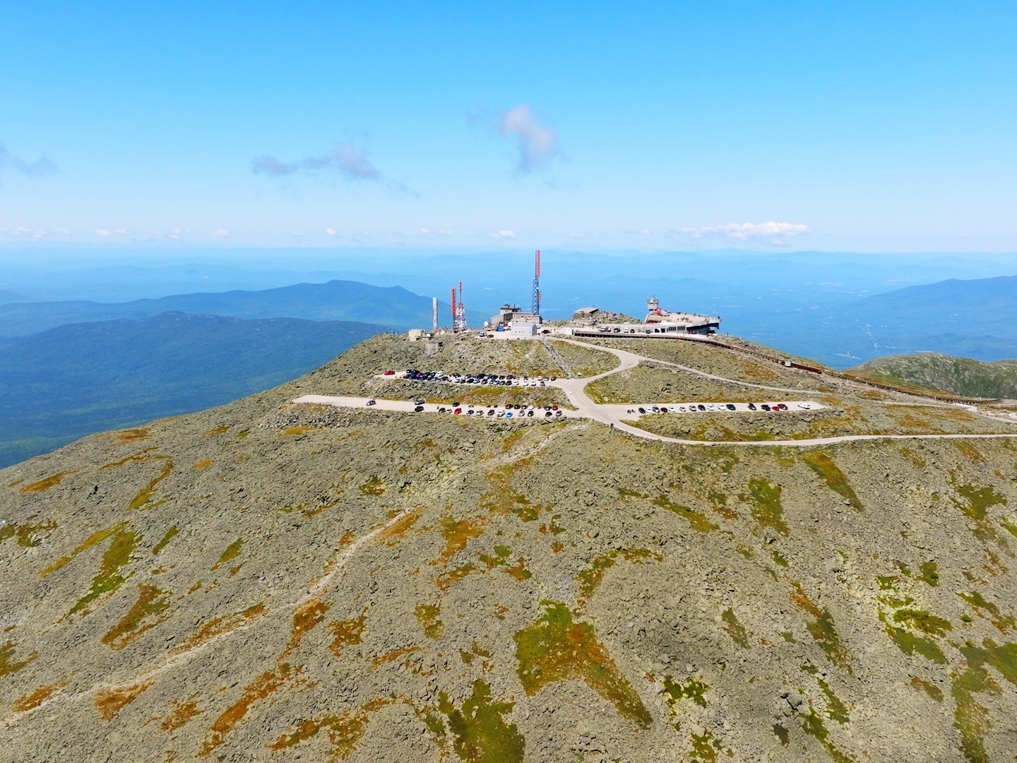 Aerial view of a rocky mountain summit with a building and parked cars under a clear blue sky.