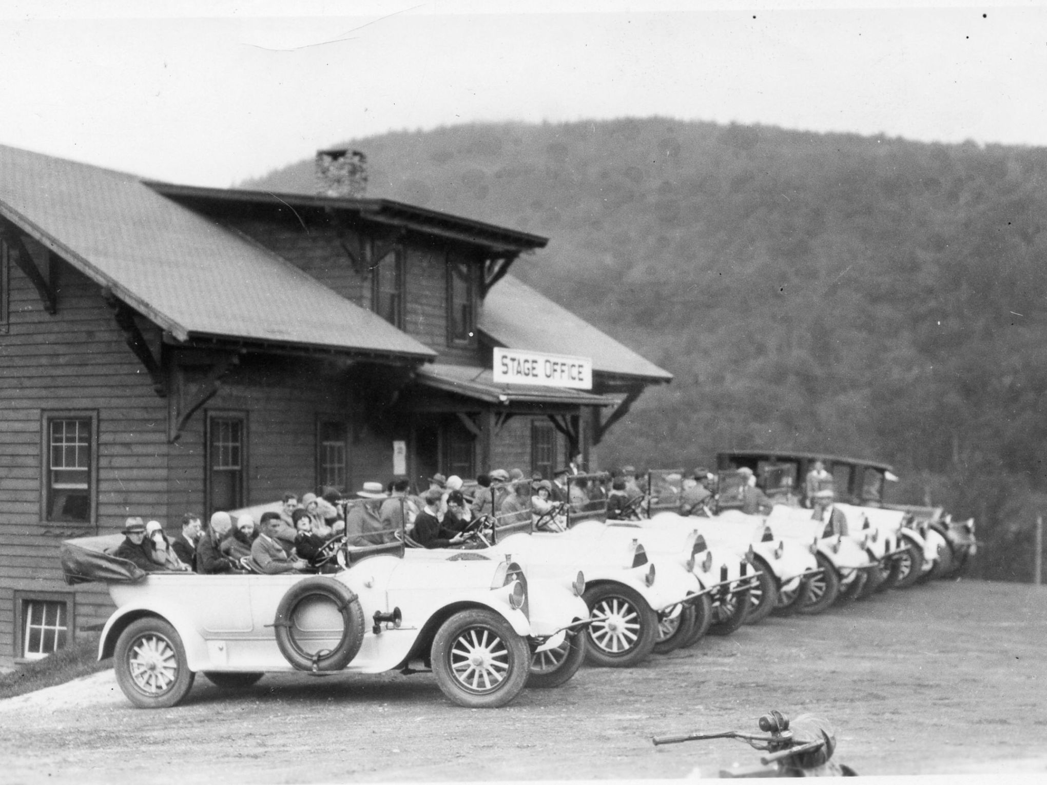 Vintage cars with passengers lined up in front of a building labeled 'Stage Office' in a rural area.
