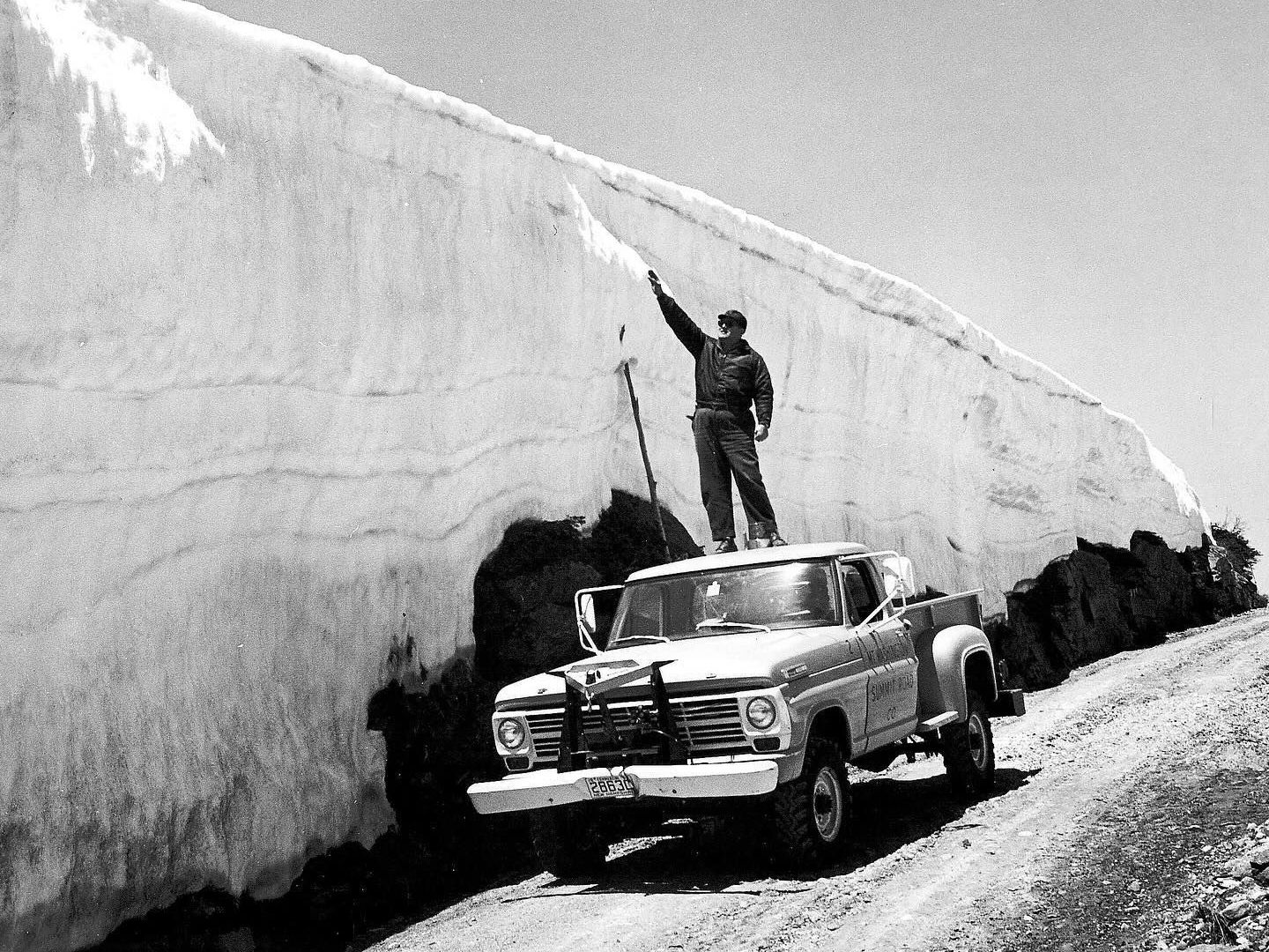 Man on truck measuring tall snowbank with stick on a dirt road.