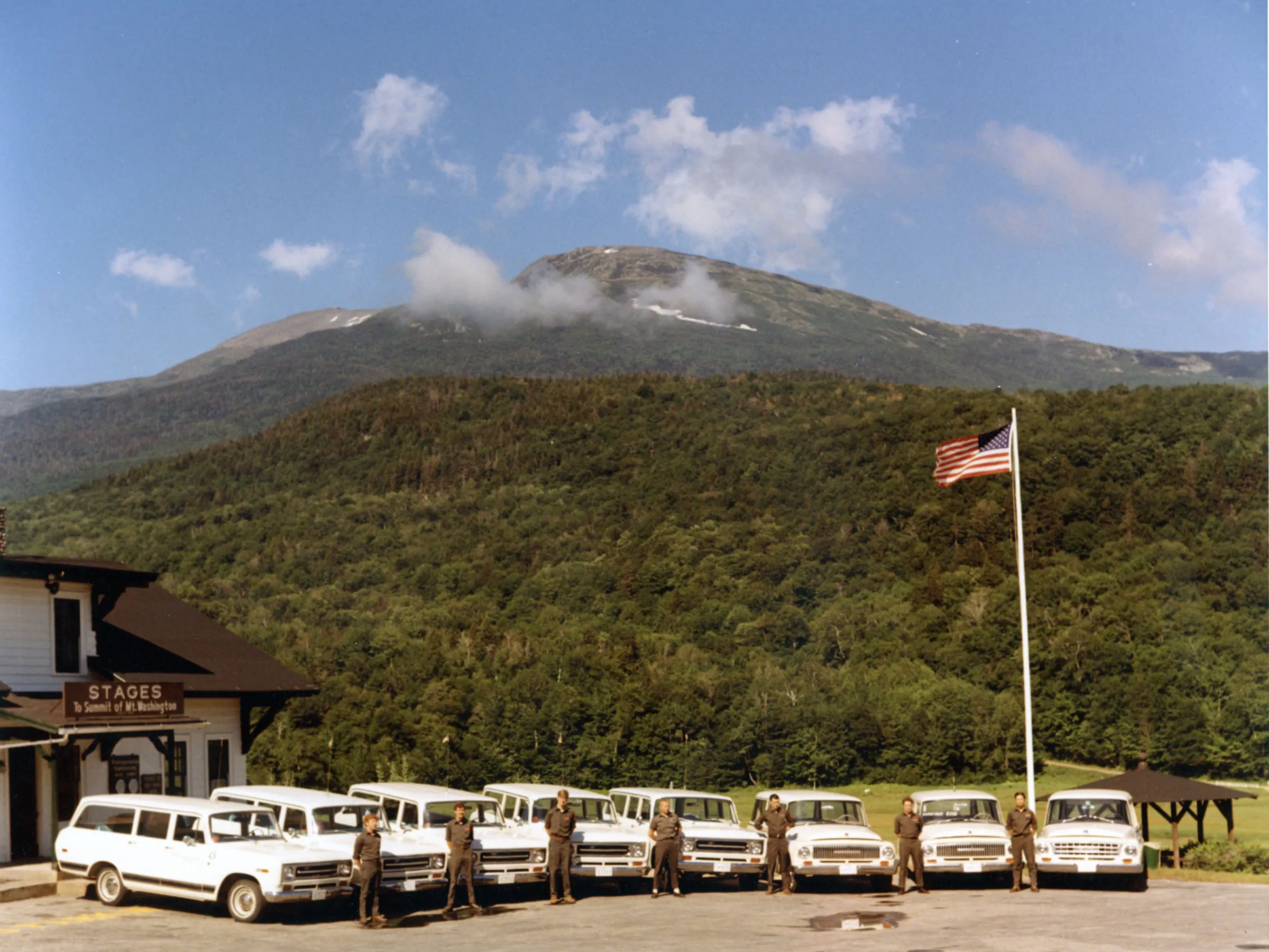 Several white vans lined up with people in uniform, American flag, and scenic mountain background.