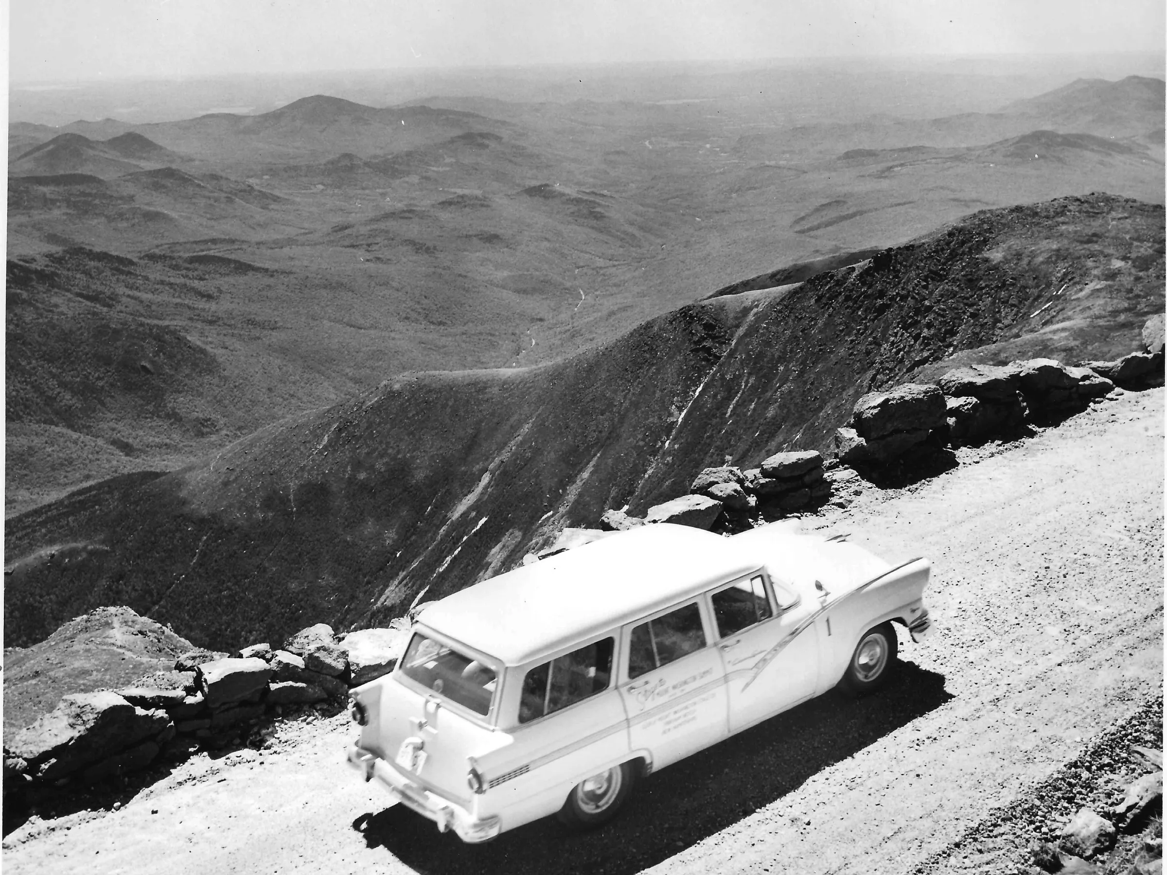 Car on a mountain road with a vast mountainous landscape in the background.