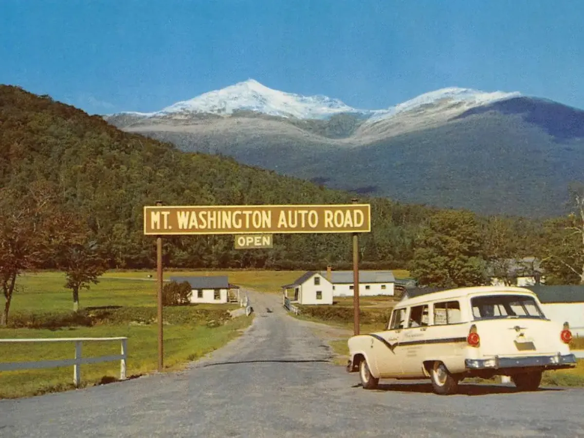 Car by Mt. Washington Auto Road sign with mountain in background on a sunny day.