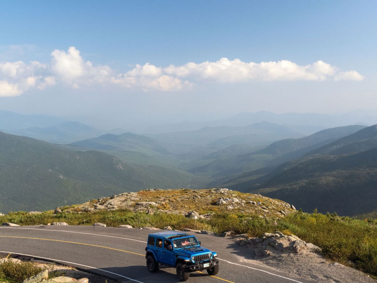 Blue Jeep driving on a mountain road under a clear blue sky with distant hills.