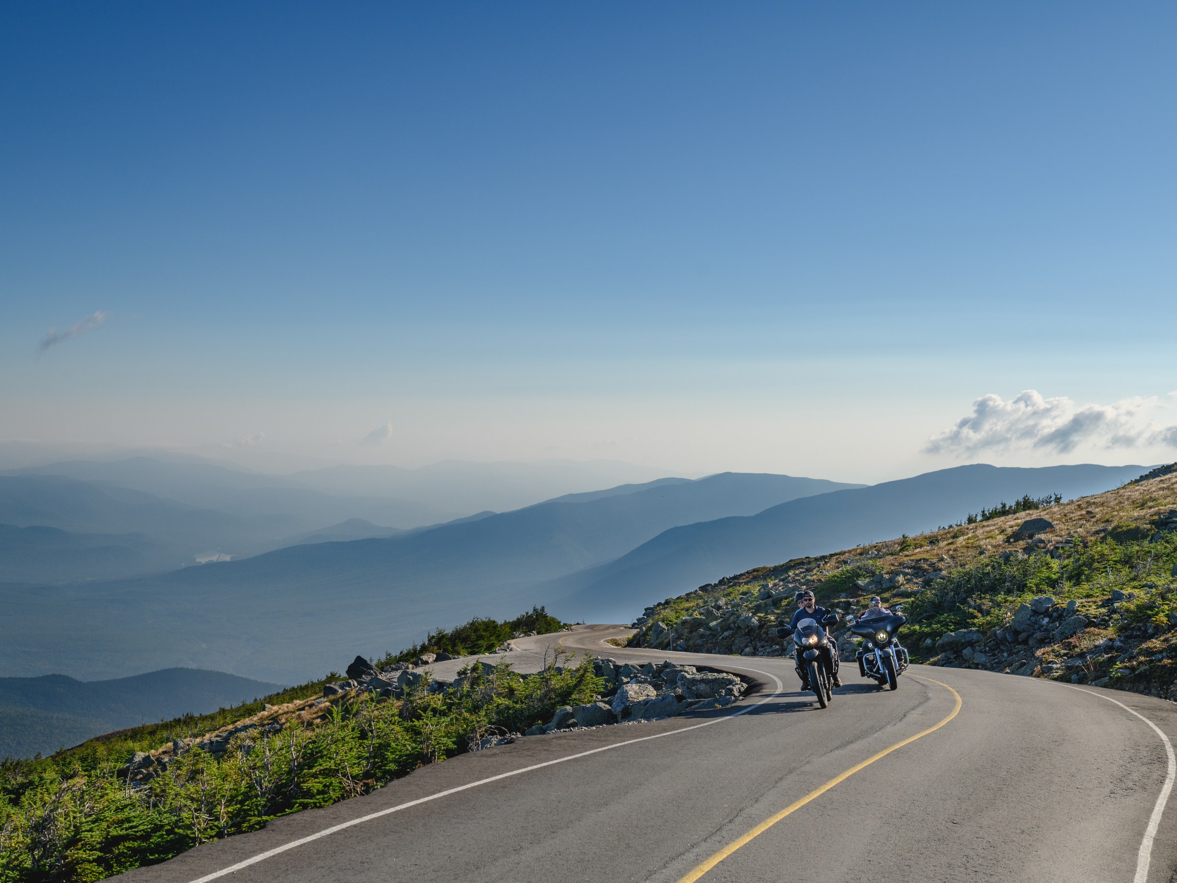 Two motorcyclists on a winding mountain road with scenic valley view under blue sky.