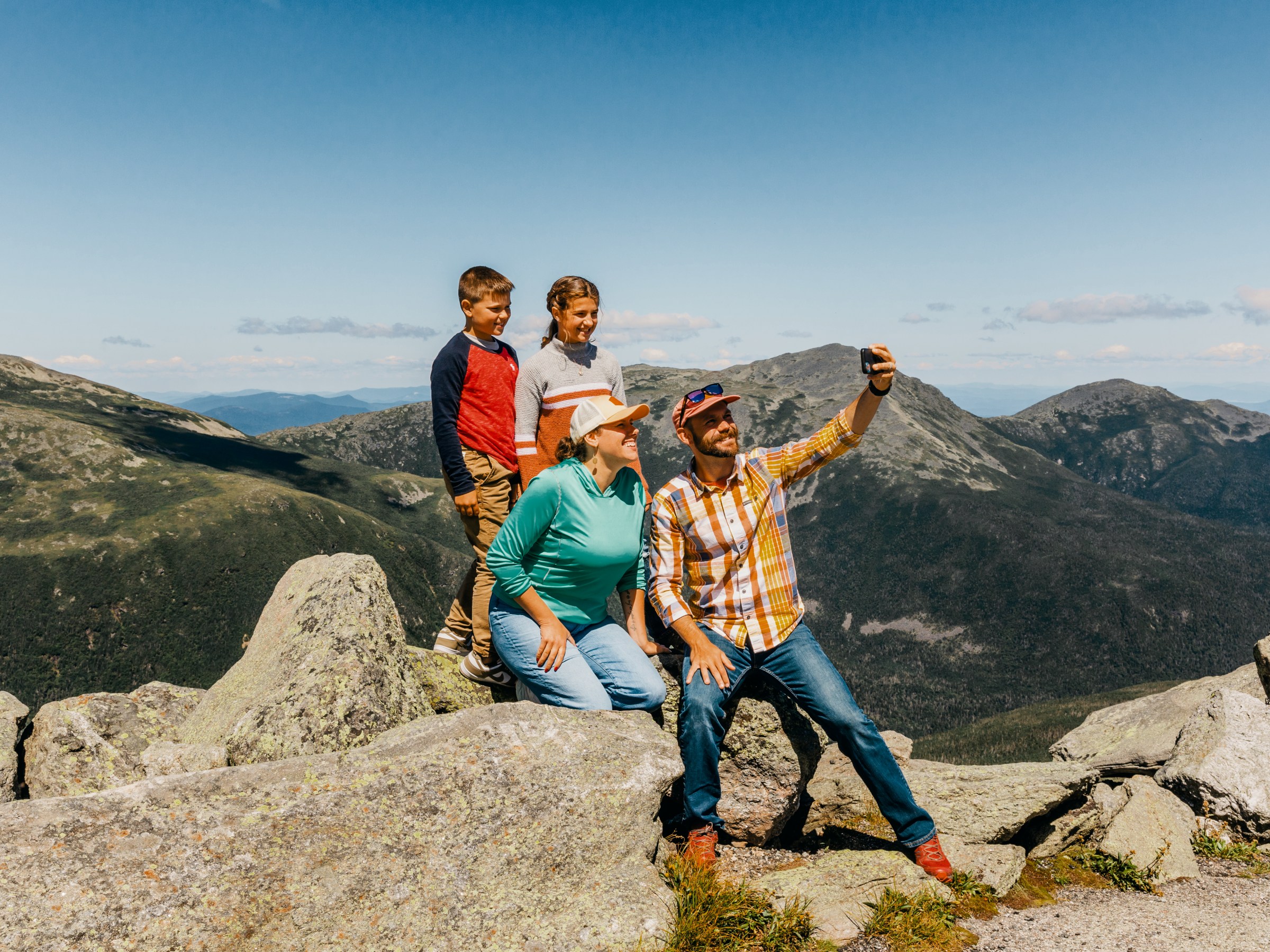 Family taking selfie on rocky mountain summit with clear blue sky.