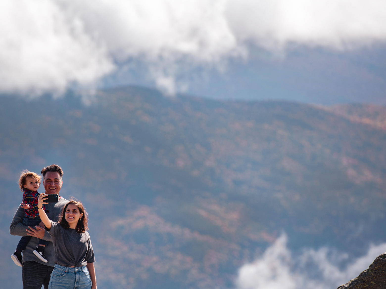 Family of three smiling in front of mountainous landscape with clouds.
